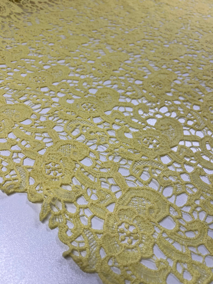 A close-up of the delicate, lightweight Anglaise Lace - Limelight from Super Cheap Fabrics reveals an intricate floral and geometric pattern. The 100% polyester fabric is spread out on a flat surface, showcasing its detailed design and edges against a neutral-colored background.