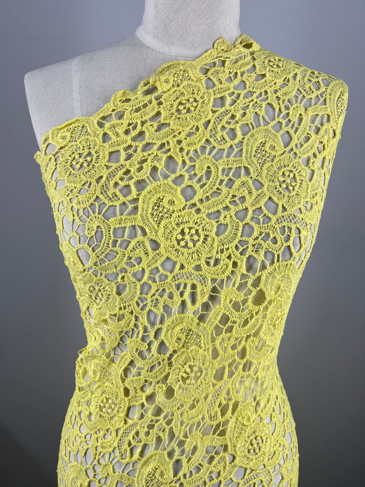 A mannequin is draped with the intricately patterned Anglaise Lace - Limelight from Super Cheap Fabrics. The 100% polyester lace, featuring floral and circular motifs, creates a visually delicate and detailed design. This lightweight lace fabric covers the mannequin asymmetrically, highlighting its texture and craftsmanship.