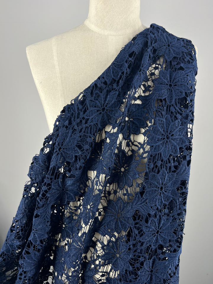 A mannequin draped in Super Cheap Fabrics' Anglaise Lace - Bloom - 120cm, an intricately crocheted lace fabric in dark blue, featuring a delicate floral pattern. Perfect for bridal wear or home décor, the lace showcases an open weave design with prominent flower motifs spread across the fabric.