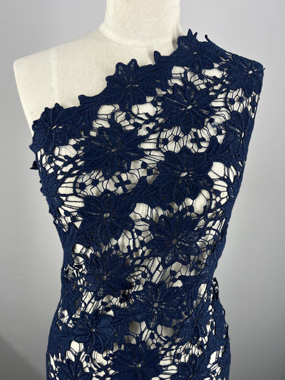 A mannequin is draped in Anglaise Lace - Bloom - 120cm from Super Cheap Fabrics, featuring dark blue floral lace with intricate flower patterns, perfect for bridal wear. The one-shoulder design exposes sections of the mannequin, highlighting the delicate and detailed craftsmanship of the lace material.