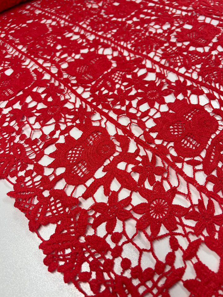 Close-up of vibrant Anglaise Lace - Red Alert - 120cm from Super Cheap Fabrics, featuring intricate floral and geometric patterns. Showcasing detailed threadwork, it highlights the fine craftsmanship perfect for bridal wear. The lace is spread flat, partially rolled on one side, making it ideal for elegant home décor projects.