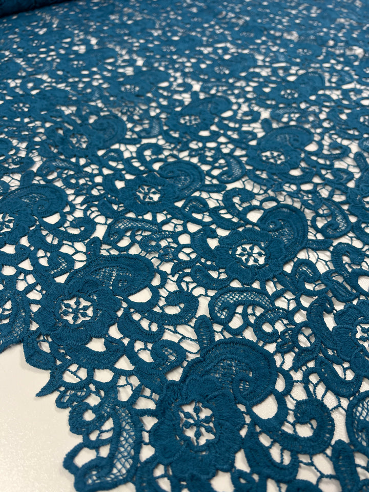 Close-up of Super Cheap Fabrics' Anglaise Lace in Teal - 120cm, showcasing intricate lace fabric with a floral and paisley pattern, ideal for home décor or bridal wear. The lace's elaborate design features delicate loops and swirling shapes, creating an elegant and airy texture on a white background.