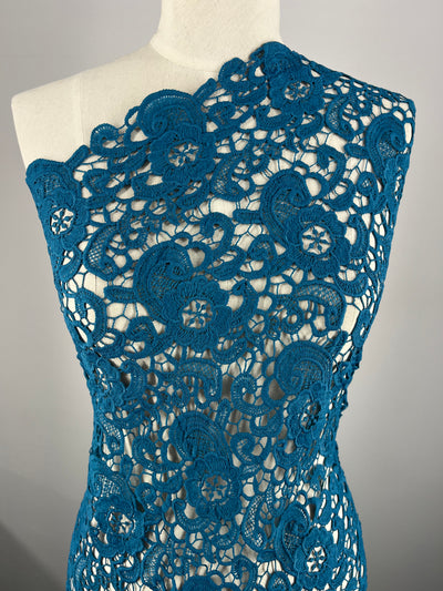 A close-up of a mannequin wearing an intricately designed dress made from Anglaise Lace - Teal - 120cm by Super Cheap Fabrics. The teal lace fabric features floral and spiral motifs, creating an elegant and detailed pattern that partially reveals the white fabric underneath. The dress has an off-shoulder style on the left side, perfect for bridal wear.