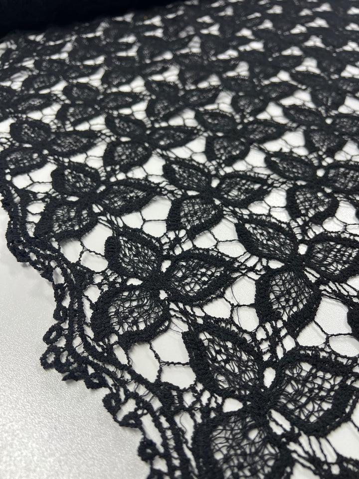 Close-up of Anglaise Lace - Black - 110cm by Super Cheap Fabrics, showcasing its intricate floral pattern. Perfect for bridal wear, the lace features delicate, interconnected flowers rendered in a fine, openwork design with scalloped edges. The background is a smooth, light-colored surface that enhances its elegance.