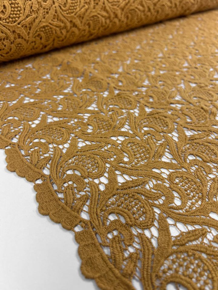 A close-up of the intricate Anglaise Lace - GOTS Cotton in Yam showcases its detailed floral and swirl patterns, making it perfect for bridal wear. The 85cm wide fabric is rolled out, revealing the ornate designs and textures with a visible scalloped edge on one side. Presented against a plain white background, this lace from Super Cheap Fabrics exudes elegance.