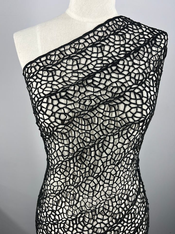 A white mannequin is adorned with Super Cheap Fabrics' Anglaise Lace in black, intricately patterned fabric that wraps diagonally across its torso. The web-like design creates a textured and elegant appearance, making it ideal for bridal wear. The background is plain and light gray.