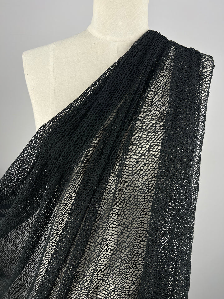 A mannequin adorned with Evening Cobble Stone Lace - Black - 150cm from Super Cheap Fabrics, which drapes loosely over its shoulder. The fabric, a lightweight sheer material made of 100% polyester, features a net-like appearance with intricate, interconnected patterns. The scene is set against a plain, neutral background.