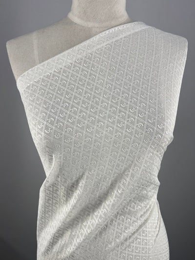 Close-up of a white, intricately patterned Stretch Mesh - Diamond - 127cm from Super Cheap Fabrics, draped on a dress form. The lightweight mesh fabric features a diamond lattice design with small, floral motifs within each diamond. The dress form is set against a grey background.