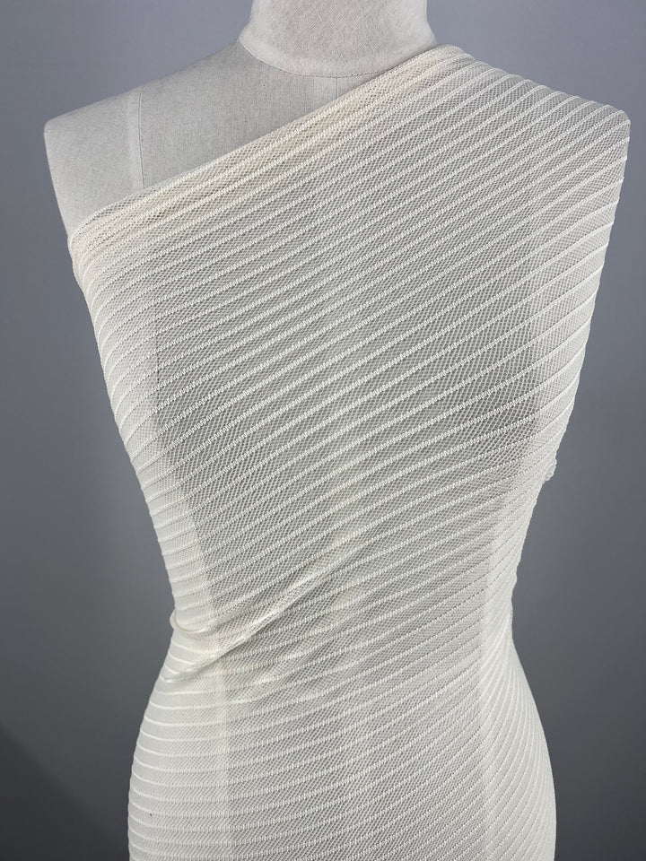 A dress form draped with Super Cheap Fabrics' Lace - Ivory Mesh - 145cm, showcasing a white, sheer, diagonal-striped material that elegantly wraps around the body with a textured appearance. This lightweight and semi-transparent fabric is made of 100% polyester, subtly revealing the mannequin underneath against a plain gray background.