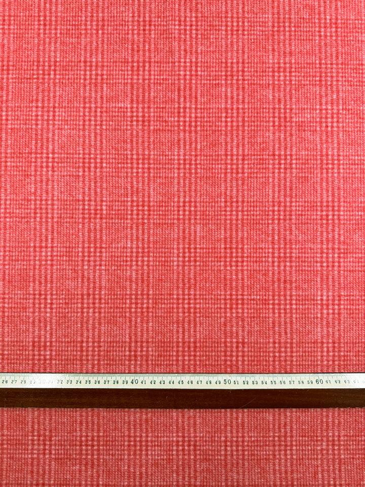 A close-up of Wool Flannel - Dubarry - 148cm by Super Cheap Fabrics, a red heavyweight fabric with a subtle grid pattern. A wooden ruler with centimeter markings is placed horizontally along the bottom of the image, showing the scale of the pattern, ideal for creating durable jackets.