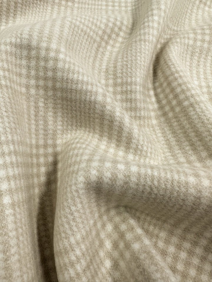 A close-up of soft, beige and cream plaid fabric, with a gentle, wavy texture. The pattern features intersecting lines forming a grid, showcasing the Super Cheap Fabrics' Wool Flannel - Parchment - 148cm heavyweight fabric’s smooth and cozy appearance perfect for jackets and coats.