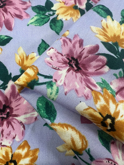 A close-up photo of Super Cheap Fabrics' Printed Rayon - Watercolour 2.0 - 145cm. The pattern showcases large pink and yellow flowers with green leaves on a light blue background. This colorful fabric is slightly folded, accentuating the vibrant and detailed print of the flowers and leaves.