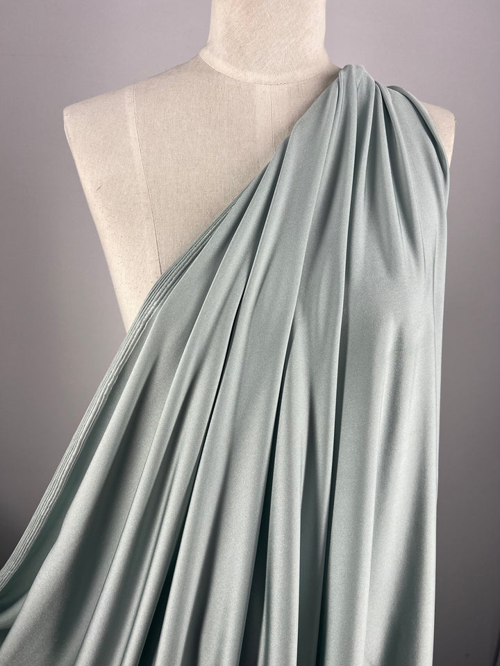 A dress form beautifully displays the Nylon Lycra - Pigeon - 150cm fabric from Super Cheap Fabrics, highlighting its light grey-blue hue and soft texture. The swimwear material elegantly flows from the shoulder, showcasing its smoothness and exceptional draping quality with its four-way stretch capability.