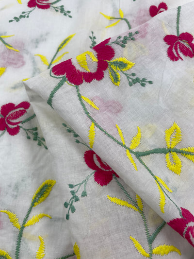 A close-up of Super Cheap Fabrics Embroidered Cotton - Anemone - 150cm featuring colorful floral embroidery. The design consists of vibrant red flowers with green stems and yellow leaves, creating an intricate pattern. The 100% cotton fabric is slightly folded in the image.