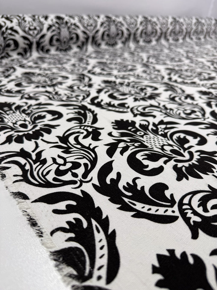 A close-up of Super Cheap Fabrics' Printed Linen Blend - Baroque Noir (145cm) shows an ornate black damask floral pattern repeating on a white background, partially unrolled to reveal its intricate design.