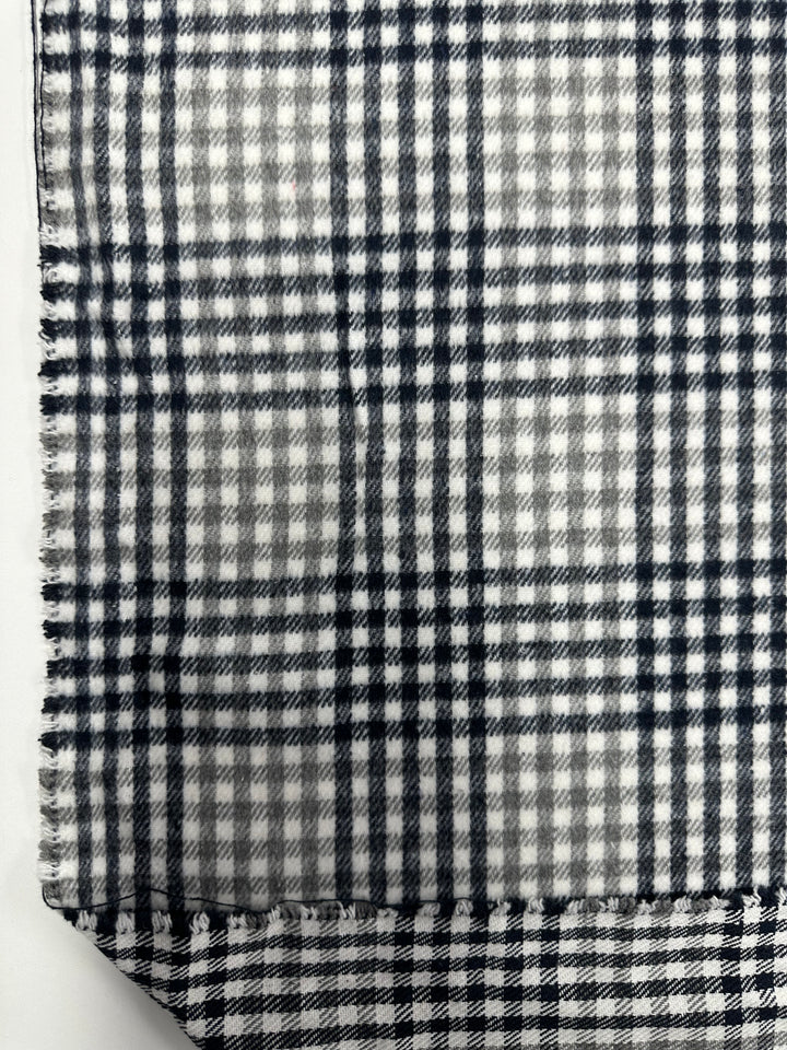 A piece of fabric with a black and white plaid pattern is shown. The **Super Cheap Fabrics Premium Brushed Plaid - Mono - 150cm**, part of our textile collection, is slightly frayed along the left edge. The bottom right corner of this medium weight fabric is folded upwards, revealing the same pattern on the reverse side.