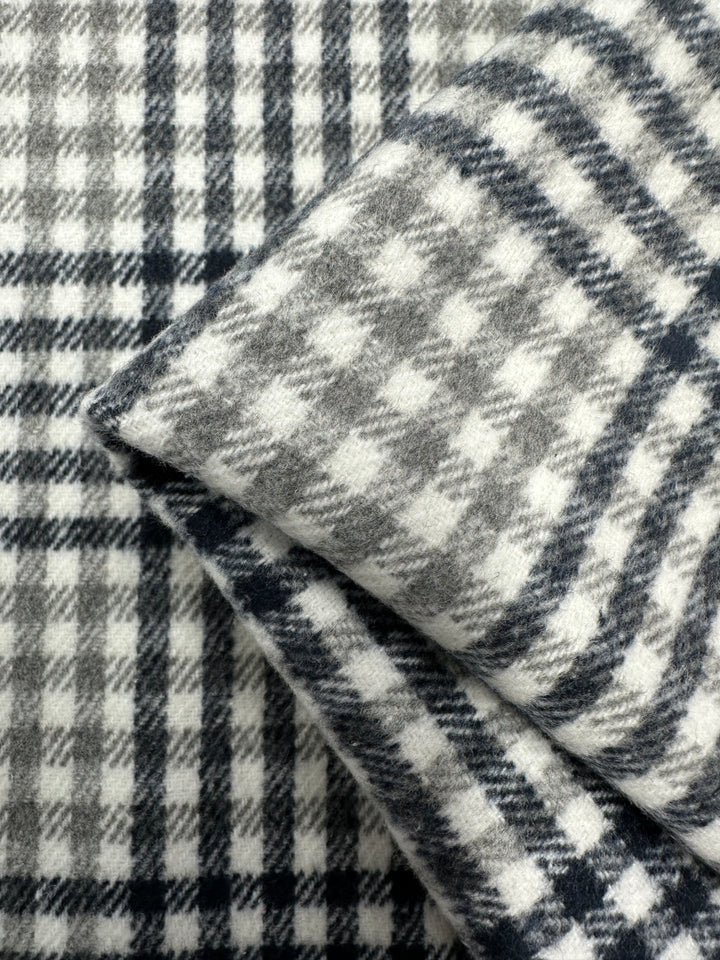 Close-up of overlapping gray and white plaid fabric from the Premium Brushed Plaid - Mono - 150cm collection by Super Cheap Fabrics. The patterns vary slightly in the arrangement and size of the checks. The texture appears soft, suggesting the medium weight fabric may be wool or a brushed cotton blend.