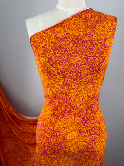 A draped mannequin displays a vibrant medium-weight fabric with an intricate orange and red pattern. The fabric cascades from the mannequin’s shoulder and down its side, showcasing the detailed design against a neutral gray background. The displayed product is Super Cheap Fabrics' Printed Lycra - Volcano Mosaic - 150cm.