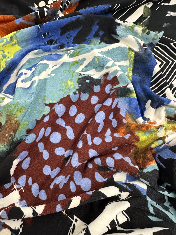 A close-up of a medium-weight fabric featuring an abstract, mixed pattern. The design includes large blue, white, and brown spots, splattered paint-like strokes in various colors, and geometric shapes. The textures are layered on this Printed Lycra - Tonal - 150cm from Super Cheap Fabrics, creating depth and visual interest.