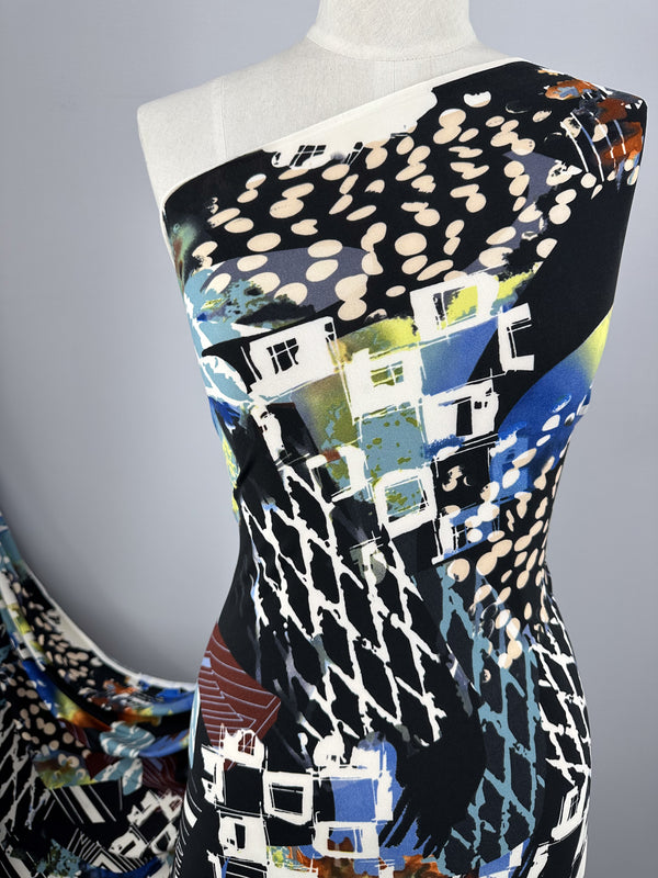 A mannequin draped in Super Cheap Fabrics' vibrant Printed Lycra - Tonal - 150cm with an abstract print. The design features a mix of geometric shapes, houses, and splashes of blue, black, white, and green. The background is a neutral grey, highlighting the fabric's intricate pattern.