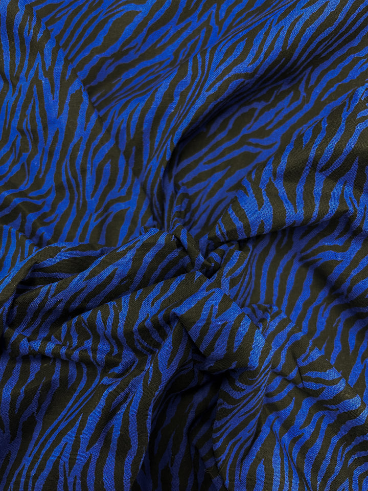 A smooth Linen Voile - Cobalt Zebra - 140cm fabric from Super Cheap Fabrics features a blue and black zebra stripe pattern. Loosely gathered in the center, it creates folds and shadows that add texture to the surface. The vibrant cobalt blue contrasts strikingly with the black background, delivering a bold and dynamic appearance ideal for home decor.