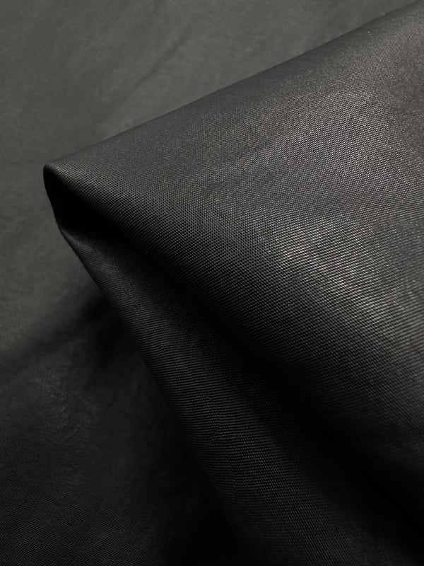 A close-up of Super Cheap Fabrics' Faux Leather - Black - 145cm, showcases its high-end, sleek, and glossy texture with a visible diagonal fold. The slightly reflective finish suggests a synthetic or coated medium-weight material.