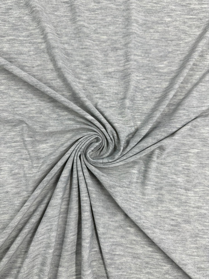 A close-up image of light gray fabric with a soft, smooth texture. The lightweight *Super Cheap Fabrics' Jersey - Grey - 170cm* is arranged in a swirl pattern, creating gentle folds and ripples radiating from the center. The overall appearance suggests a pliable and comfortable material, perfect for a stylish grey jumpsuit.