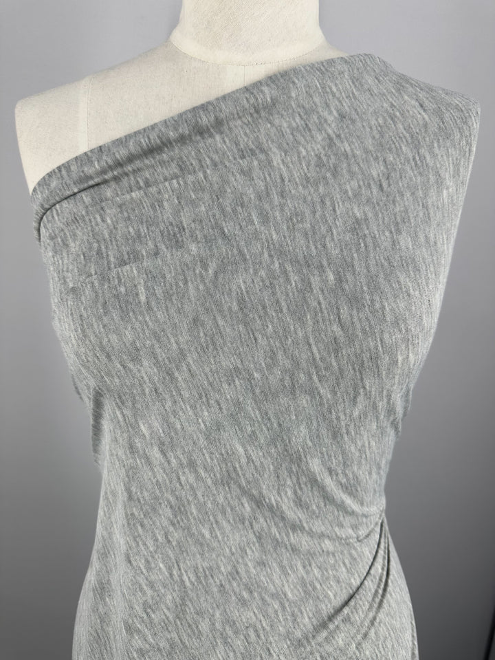 A grey, one-shoulder dress crafted from lightweight jersey fabric is displayed on a mannequin. The dress features a textured, heathered fabric that drapes smoothly over the mannequin's form, with a slight gather near the bottom right. This fabric is known as Jersey - Grey  - 170cm by Super Cheap Fabrics. The background is plain and gray.