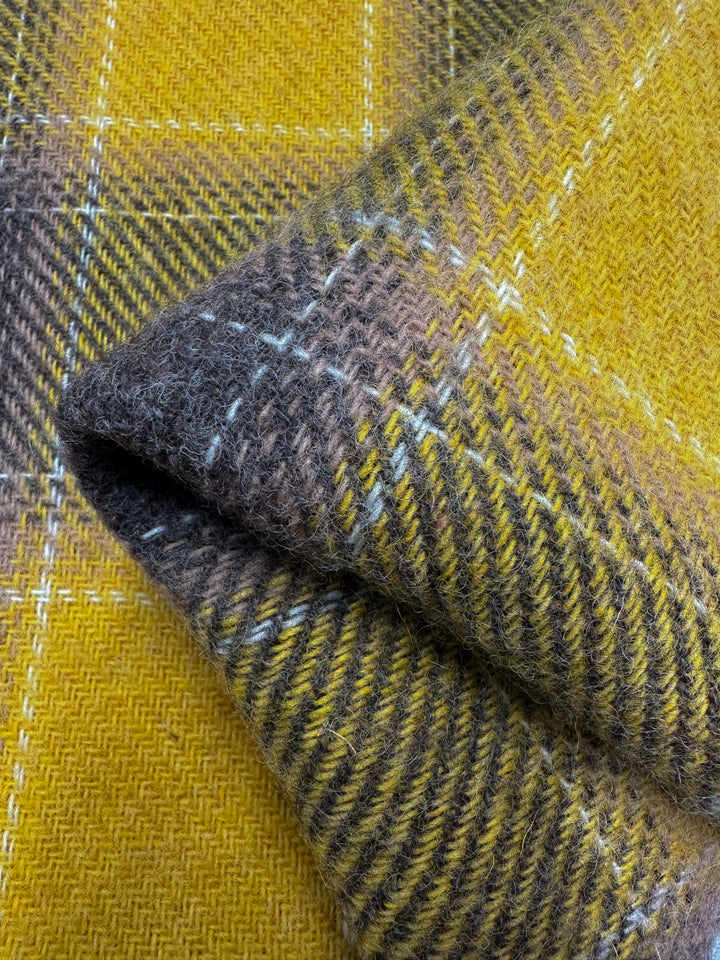 Close-up of a folded piece of fabric with a plaid pattern. The material, Virgin Wool - Spruzzo by Super Cheap Fabrics, showcases a combination of yellow and brown hues in a traditional tartan design. The heavy-weight wool fabric features visible stitching and texture, appearing soft and ideal for overcoats made from virgin wool.