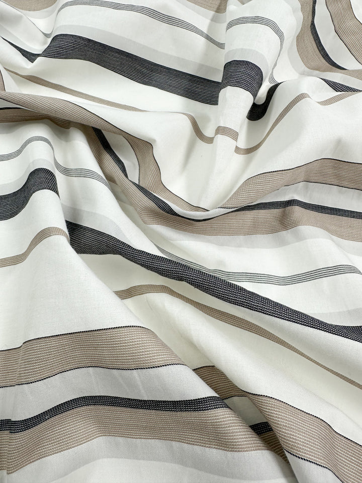 A close-up image of draped, lightweight fabric featuring horizontal stripes in beige, black, and white. The Cotton Voile - Creamy Nimbus from Super Cheap Fabrics appears smooth and soft with gentle folds and a subtle sheen, creating an elegant and stylish pattern.