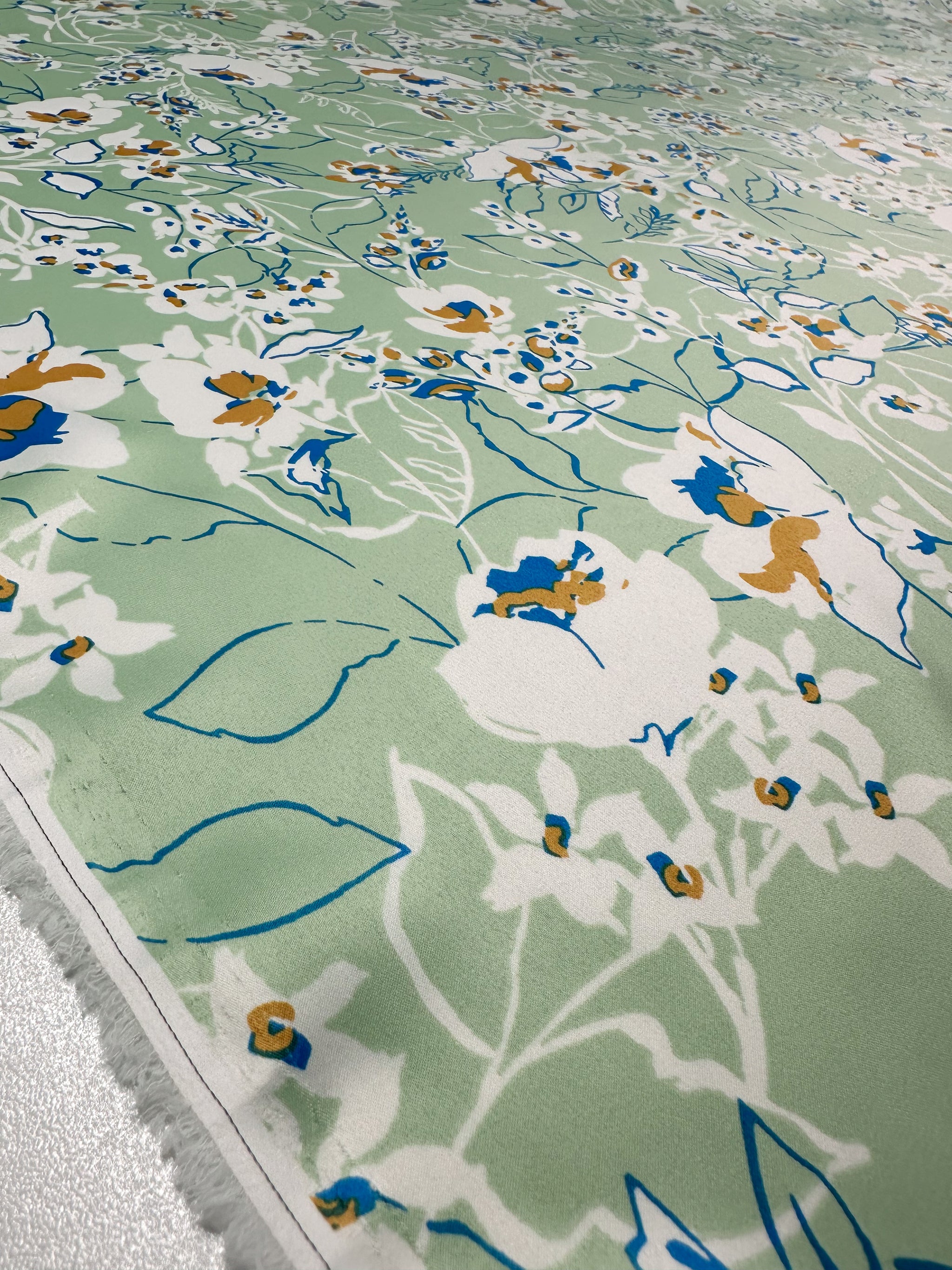 The Printed Stretch Satin - Green Sakura by Super Cheap Fabrics offers a close-up of lightweight fabric in light green, showcasing an intricate floral pattern in white, blue, and orange with various leaves and flowers for a vibrant look. Width: 150cm.