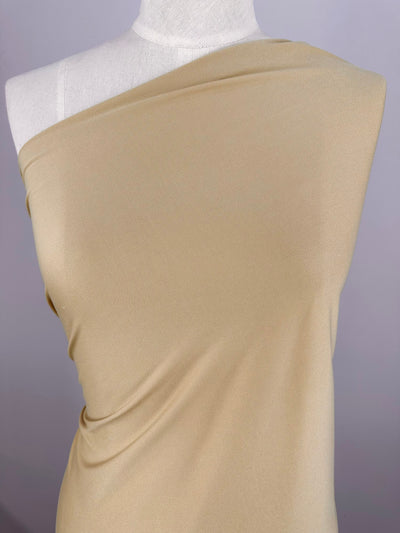 A beige, lightweight fabric draped over a dress form, showcasing its smooth texture and subtle sheen. Made from 100% polyester, the Interlock - Sand - 150cm by Super Cheap Fabrics is displayed in a way that accentuates its drape and flow, suggesting its suitability for garment creation. The background is plain and neutral.