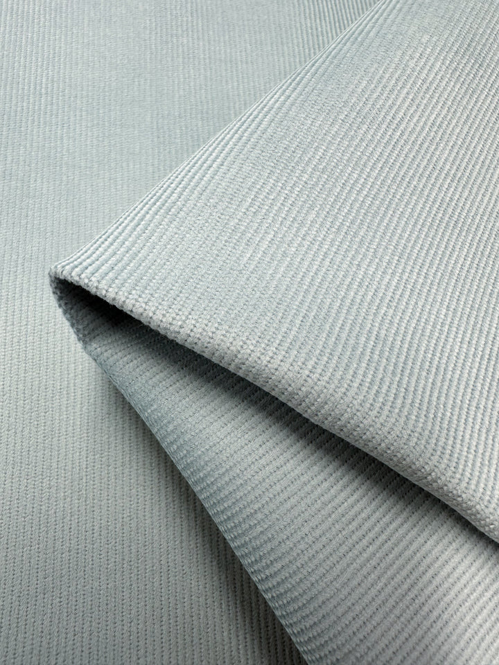 Close-up of folded Spa Blue Micro Wale Corduroy fabric by Super Cheap Fabrics, showcasing its textured, ribbed pattern. The fabric appears smooth and durable, with shallow ridges creating a subtle striped effect. The folds highlight the fabric's flexibility and thickness, making it ideal for home decor or cozy winter clothing at 145cm width.