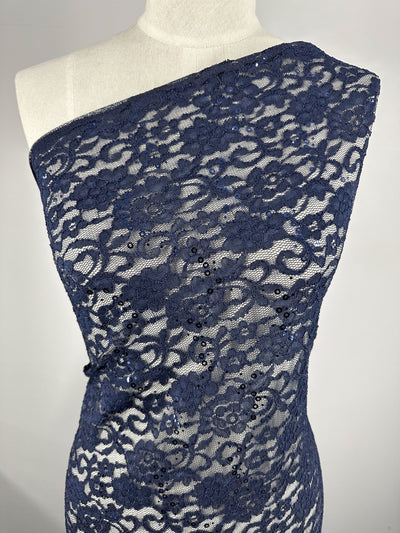 A dress form adorned with Super Cheap Fabrics' Evening Lace - Night Sky, a dark blue, one-shoulder floral lace fabric made from 100% polyester. The intricate, lightweight material showcases a detailed flower pattern embellished with sequins for added sparkle. Its sheer quality stands in striking contrast against the plain white wall background.