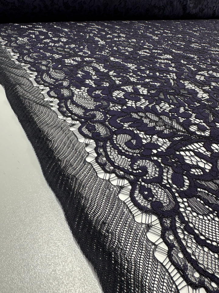 A close-up image of Super Cheap Fabrics' Evening Lace - Letita - 148cm, laid on a flat surface. The dark navy sheer material features an intricate floral pattern with scalloped edges, displaying detailed craftsmanship and texture. The neutral light background accentuates the elegance and lightweight quality of this exquisite lace design.