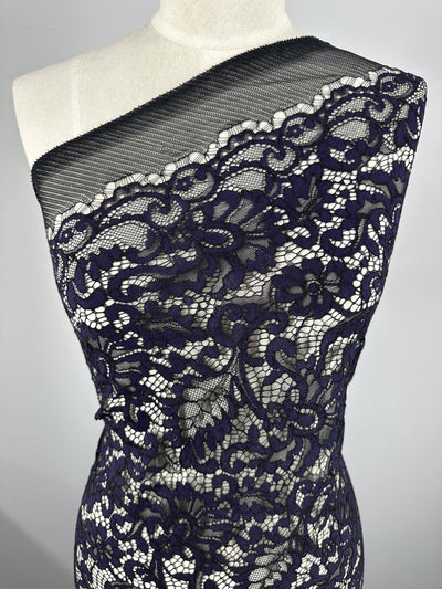 A dress form adorned with Super Cheap Fabrics' Evening Lace - Letita - 148cm, featuring a dark navy floral lace with intricate flower and vine patterns on a sheer mesh base. This lightweight fabric is arranged asymmetrically, covering one shoulder and draping diagonally across the torso.