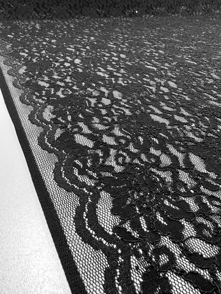 A close-up black and white image of Super Cheap Fabrics' "Evening Lace - Twinkle - 133cm" showcases an intricately detailed floral pattern. This polyester lace fabric is laid out flat, revealing its delicate texture that extends into the background, hinting at its potential use in lightweight clothing or sheer black fabric designs.