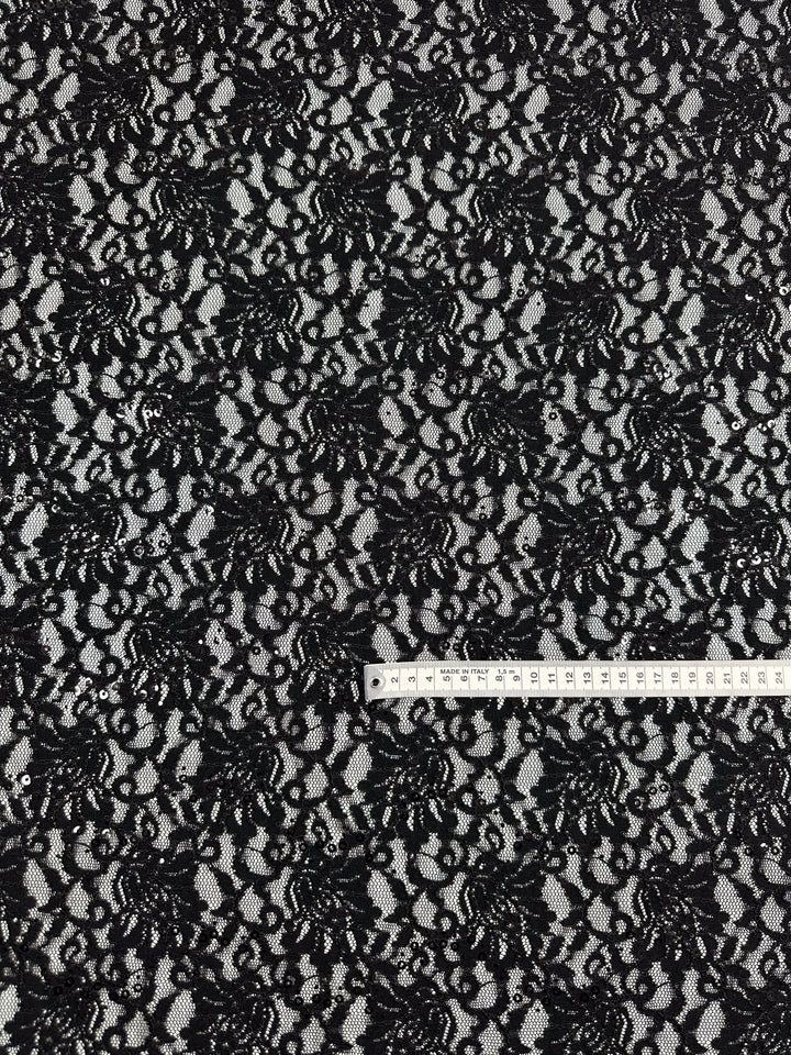 A piece of Super Cheap Fabrics' Evening Lace - Twinkle - 133cm, featuring intricate floral patterns, is spread out. A white measuring tape displaying both inches and centimeters is placed horizontally across the bottom part of the black sheer fabric.