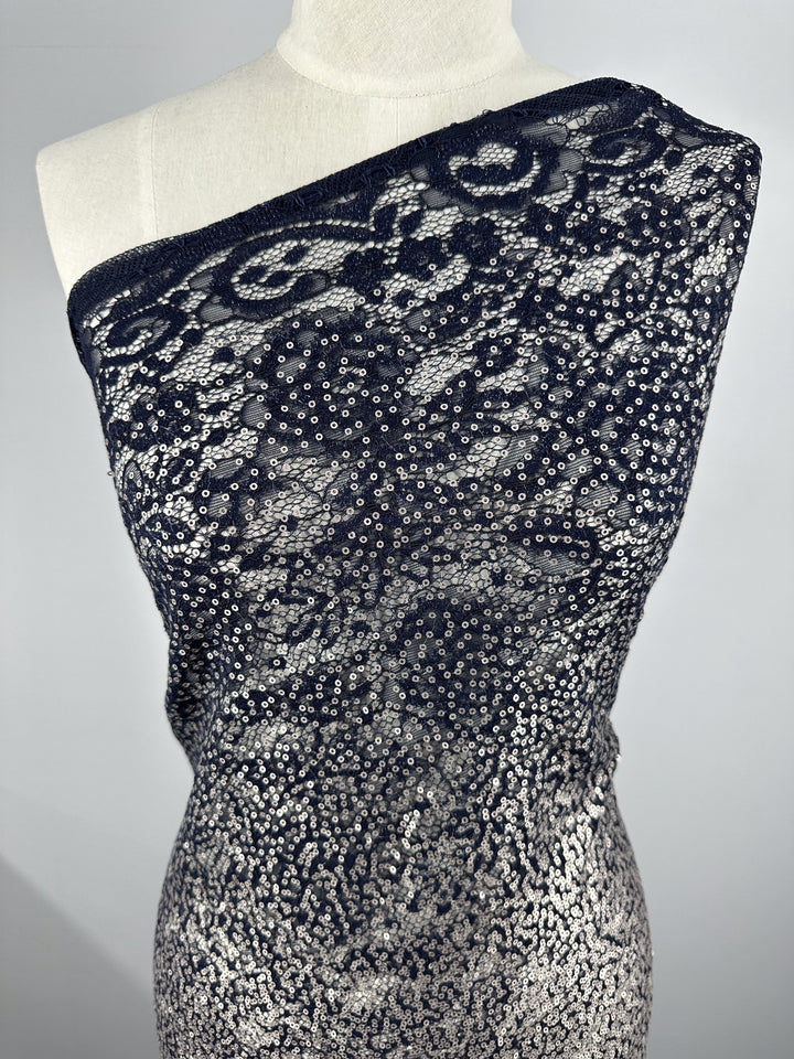 A close-up shot of a mannequin wearing the Evening Lace - Holy Moly - 145cm from Super Cheap Fabrics, an off-the-shoulder dress adorned with intricate black lace and silver sequins. The sheer fabric dress features a floral pattern and sparkly details, creating a sophisticated and elegant look.