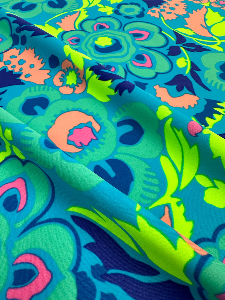 A close-up of the Super Cheap Fabrics' Printed Nylon Lycra - Mirage - 158cm, showcasing a high-quality print with an abstract floral pattern in vibrant shades of blue, green, pink, and yellow. The light to medium weight fabric features layered, swirled shapes reminiscent of flowers and leaves, slightly folded to highlight its texture and dynamic design.