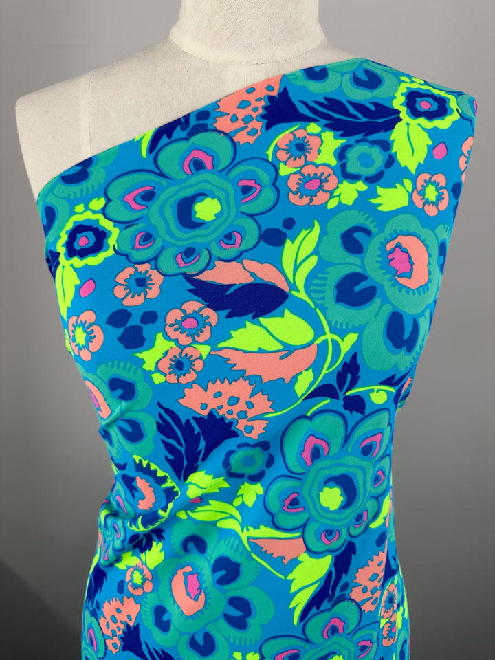 A mannequin is dressed in a one-shoulder garment featuring the vibrant and colorful floral print of Super Cheap Fabrics' Printed Nylon Lycra - Mirage - 158cm. The high-quality print includes large flowers in shades of blue, green, and pink against a predominantly blue background. The light to medium weight fabric makes it perfect for stylish swimwear.