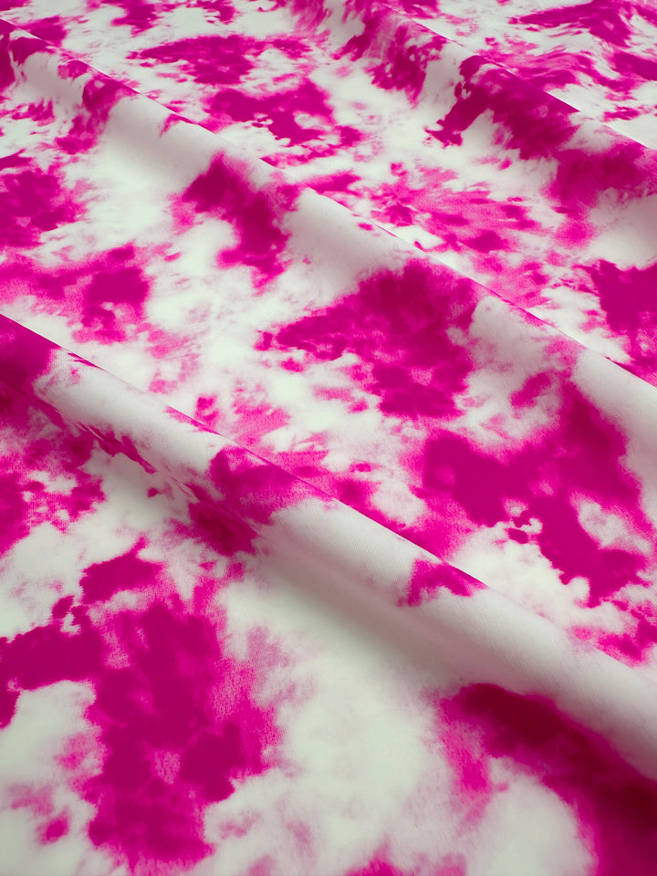 A close-up view of the Printed Nylon Lycra - Bandhani from Super Cheap Fabrics showcases a soft, lightweight material with a vibrant pink and white tie-dye pattern. The fabric is slightly wrinkled, adding shadows and texture that highlight its high-quality print. Irregular splotches and swirls blend seamlessly, making it ideal for versatile multi-use clothing.