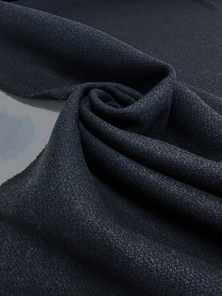 A close-up of the Wool Tweed - Vulcan fabric by Super Cheap Fabrics reveals its dark hue swirled into a spiral. The 150cm material, ideal for outer coats, has a thick appearance with a subtle shine and visible weave, adding depth and detail.