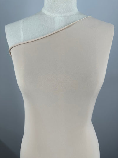 A dress form mannequin is adorned with a beige, medium weight fabric one-shoulder garment. The ITY Knit - Nude - 150cm from Super Cheap Fabrics has a smooth and slightly shiny texture, extending down to an unseen part of the mannequin. The background is evenly lit with a neutral, gradient effect.