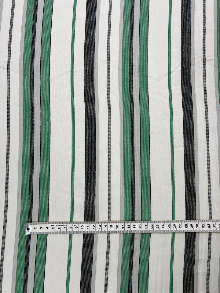 A fabric from our Designer Cotton Range, specifically the Super Cheap Fabrics Designer Cotton - Green Rock Candy - 145cm, features vertical stripes in black, gray, and green on a white background. A measuring tape is placed horizontally at the bottom of the image, showing measurements in centimeters from 0 to 34, highlighting our commitment to sustainable deadstock materials.