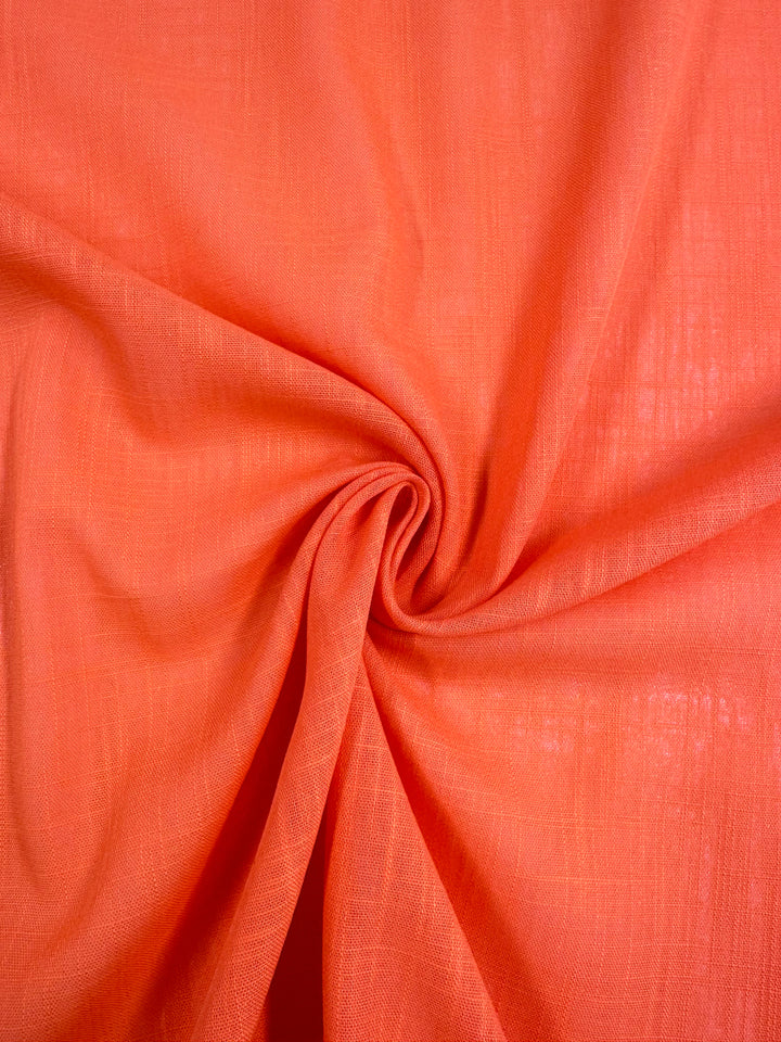 A close-up image of the bright orange Linen Blend - Camellia from Super Cheap Fabrics, showcasing its soft texture. The fabric, a lightweight linen cotton blend with a subtly visible weave pattern, is elegantly arranged in gentle folds that converge at the center in a twist, creating a dynamic visual effect.