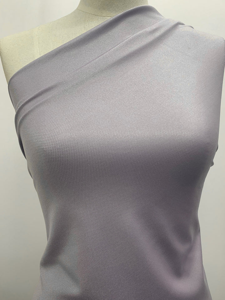 A mannequin showcases the Super Cheap Fabrics' Nylon Lycra Lilac Scale, a 140cm wide single-shoulder top. This piece comes in a gentle lilac hue with an understated textured design, crafted from soft nylon lycra fabric that subtly shimmers in the light.