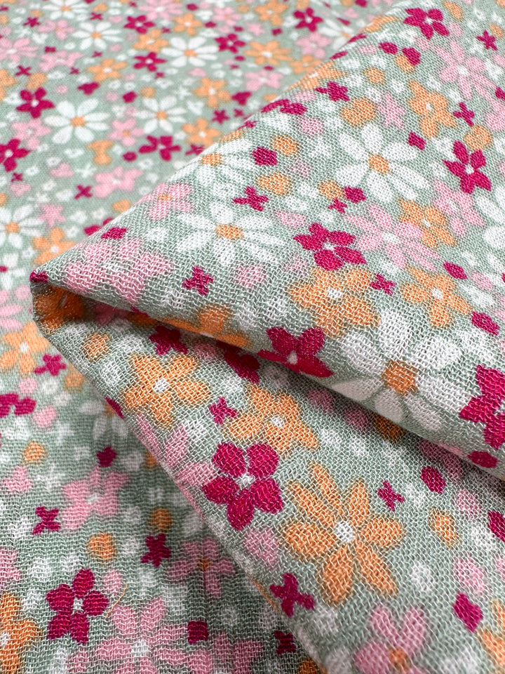 Close-up of the Soft Cotton - Cluster fabric by Super Cheap Fabrics, featuring a breathable design with a floral pattern. It showcases small pink, orange, and white flowers on a light green background. The fabric is folded to illustrate its vivid and intricate design, making it an ideal choice for summer garments.