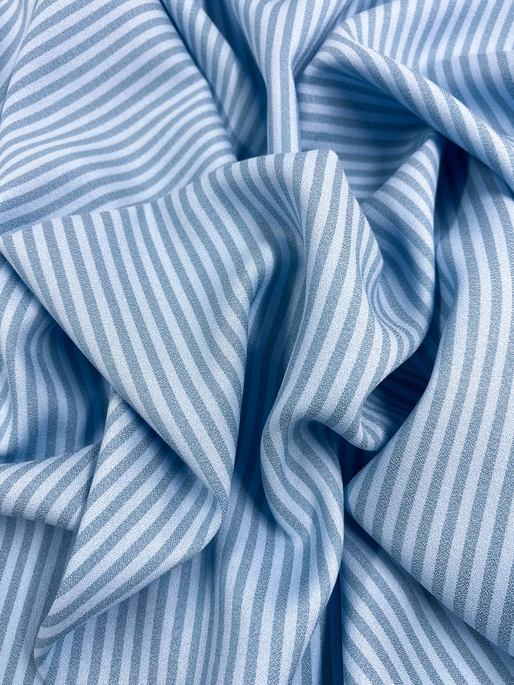 A detailed view of the Striped Crepe - Milky Blue by Super Cheap Fabrics showcases its light blue and milky blue stripes made from polyester, gently wrinkled to form soft folds and shadows. The pattern features evenly spaced vertical stripes, imparting an elegant touch.