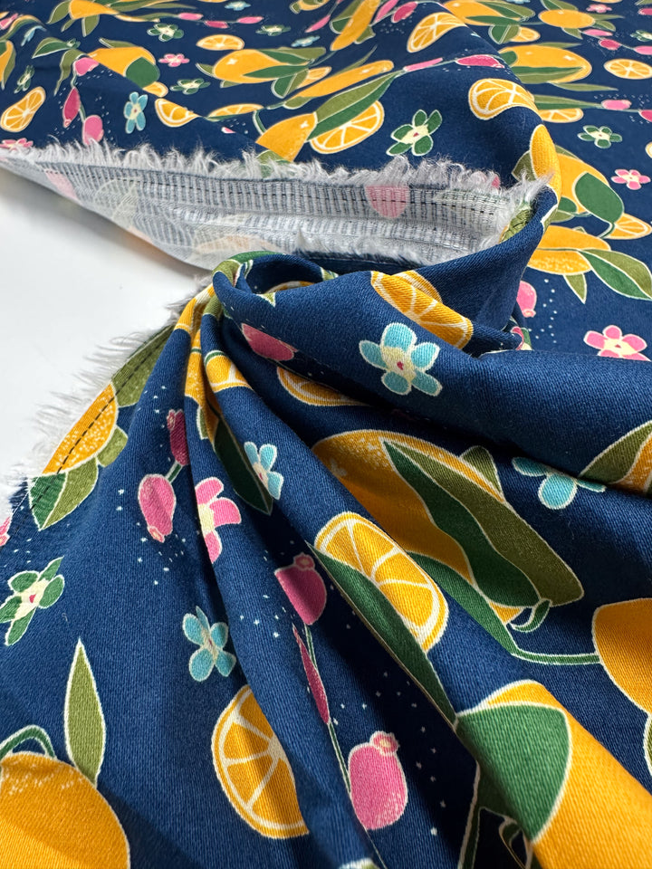 A close-up of Super Cheap Fabrics' Cotton Sateen - OJ (145cm), a medium weight cotton with vibrant oranges, lemons, flowers, and leaves on dark blue. The fabric is partially folded, showing the underside.